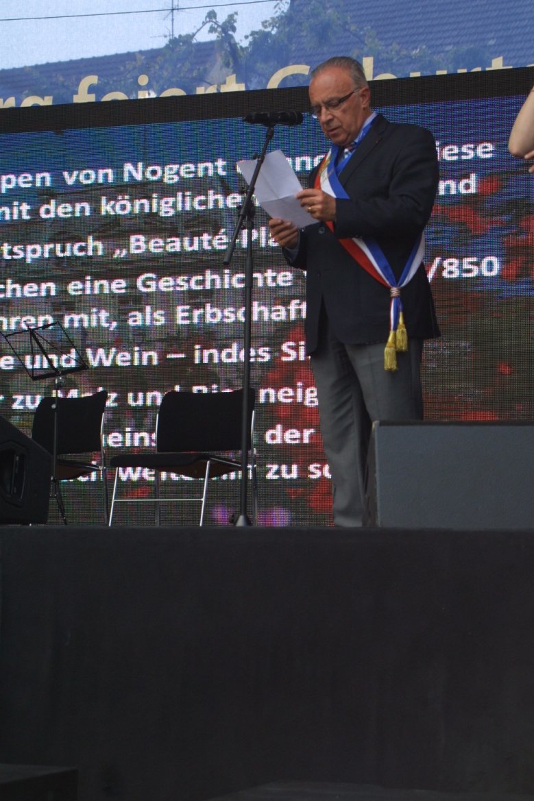 2020-07-01_jacques-martin Mann mit Trikolore steht auf einer Bühne und liest eine Rede vom Zettel ab, die im Hintergrund auf digitaler Leinwand eingeblendet wird