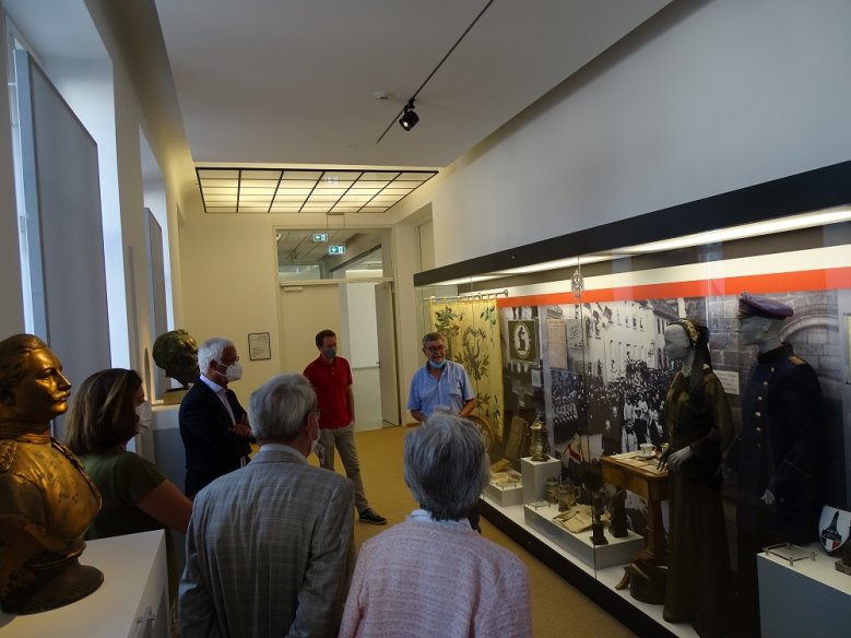 2020-06-24 Vitrine-stadtmuseum Mehrere Personen mit Mund-Nase-Bedeckung stehen vor einer Vitrine mit Ausstellungsgegenständen, ein Mann steht davor und erklärt