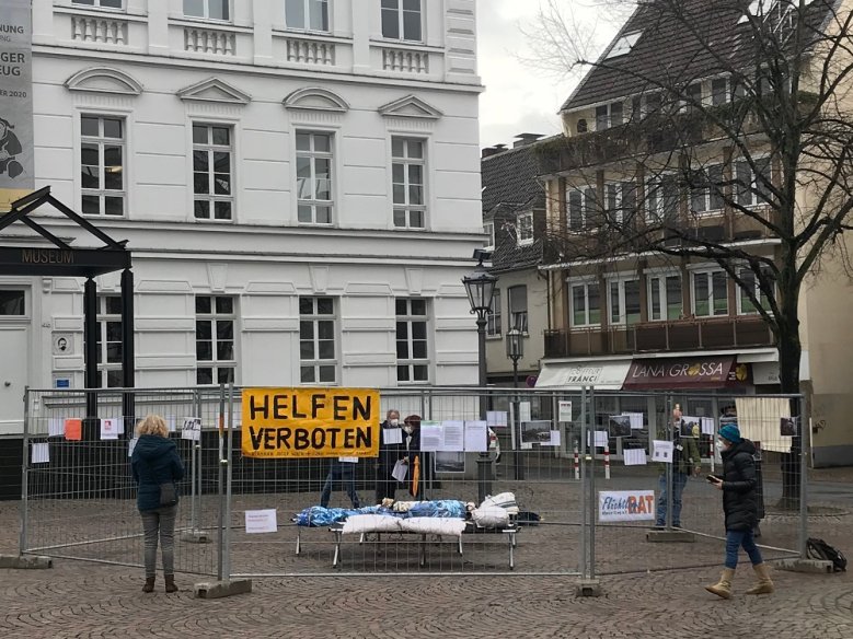 Helfen verboten Klappbetten hinter einem Bauzaun, daran ein Plakat mit der Aufschrift Helfen verboten