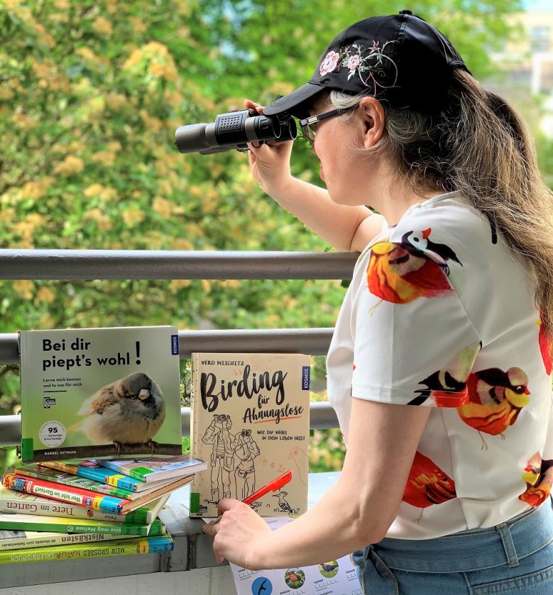 Bibliothek-Schulstunde der Gartenvögel Frau mit Fernglas blickt ins Grüne, vor ihr stehen verschiedene Vogelkunde-Bücher