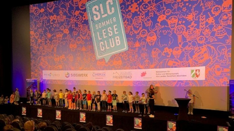 Finale SLC Kinder auf der Bühne im großen Cineplex-Saal vor dem SommerLeseClub-Logo