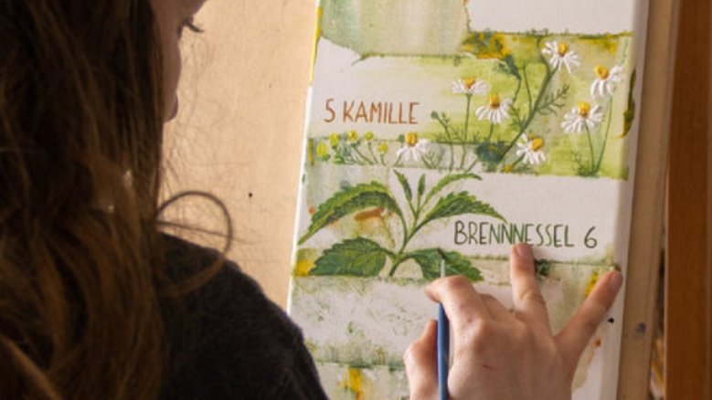 PachaPan Kräuterkompositionen Frau vor einer Leinwand, auf die sie Bilder von Kamille und Brennnessel zeichnet