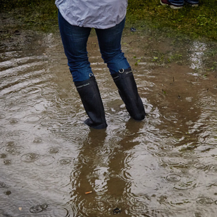 Das Bild zeigt eine überflutete Straße. Eine Frau in Stiefeln läuft durch das Wasser