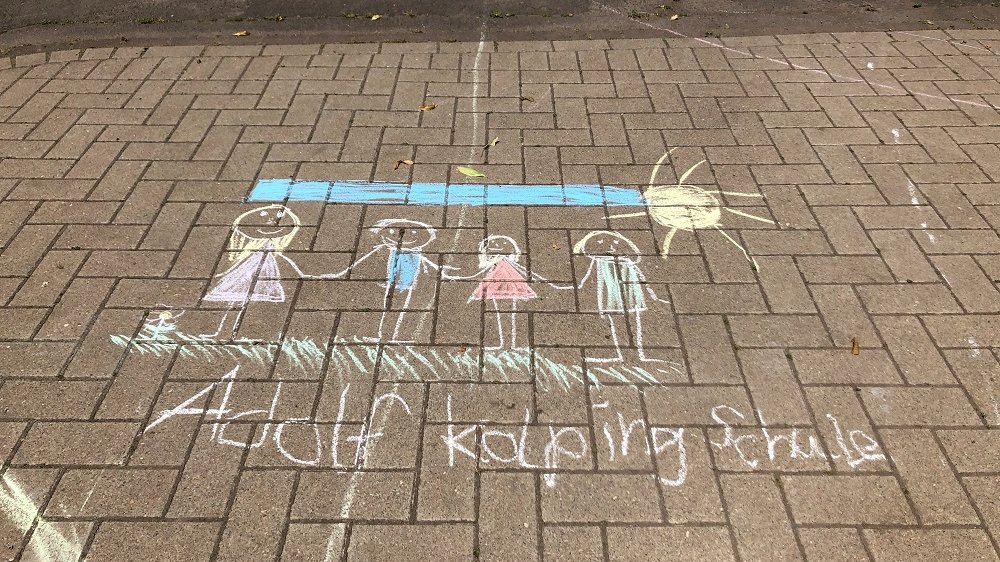 Kreidezeichnung eines Kindes, dieses zeigt vier Personen, die sich an den Händen halten, darüber ein blauer Himmel und eine Sonne, darunter der Schriftzug Adolf Kolping Schule