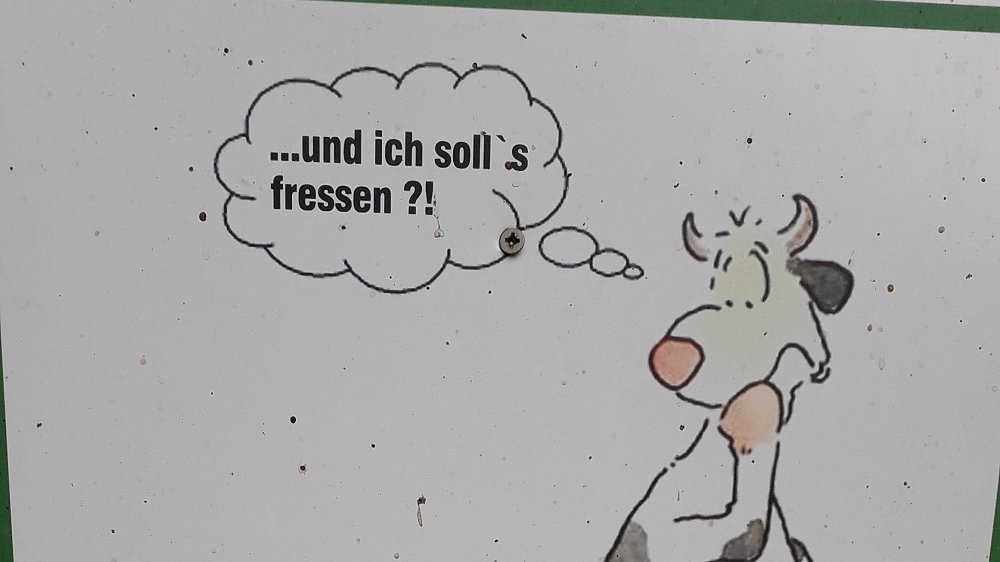 Schild mit Zeichnung eines Hundes, der neben einer stehenden Kuh auf die Wiese kotet. Die Kuh fragt sich: "... und ich soll's fressen?"