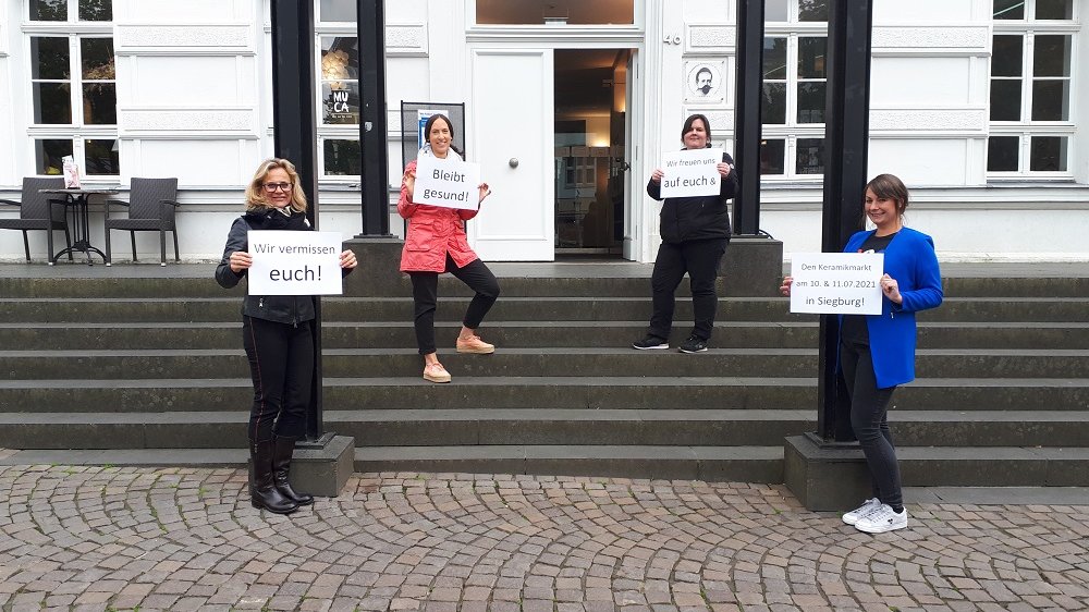 Vier Frauen stehen auf einer Treppe und halten Schilder hoch: "Wir vermissen euch! Bleibt gesund! Wir freuen uns auf euch & den Keramikmarkt am 10. & 11.07.2021 in Siegburg!"