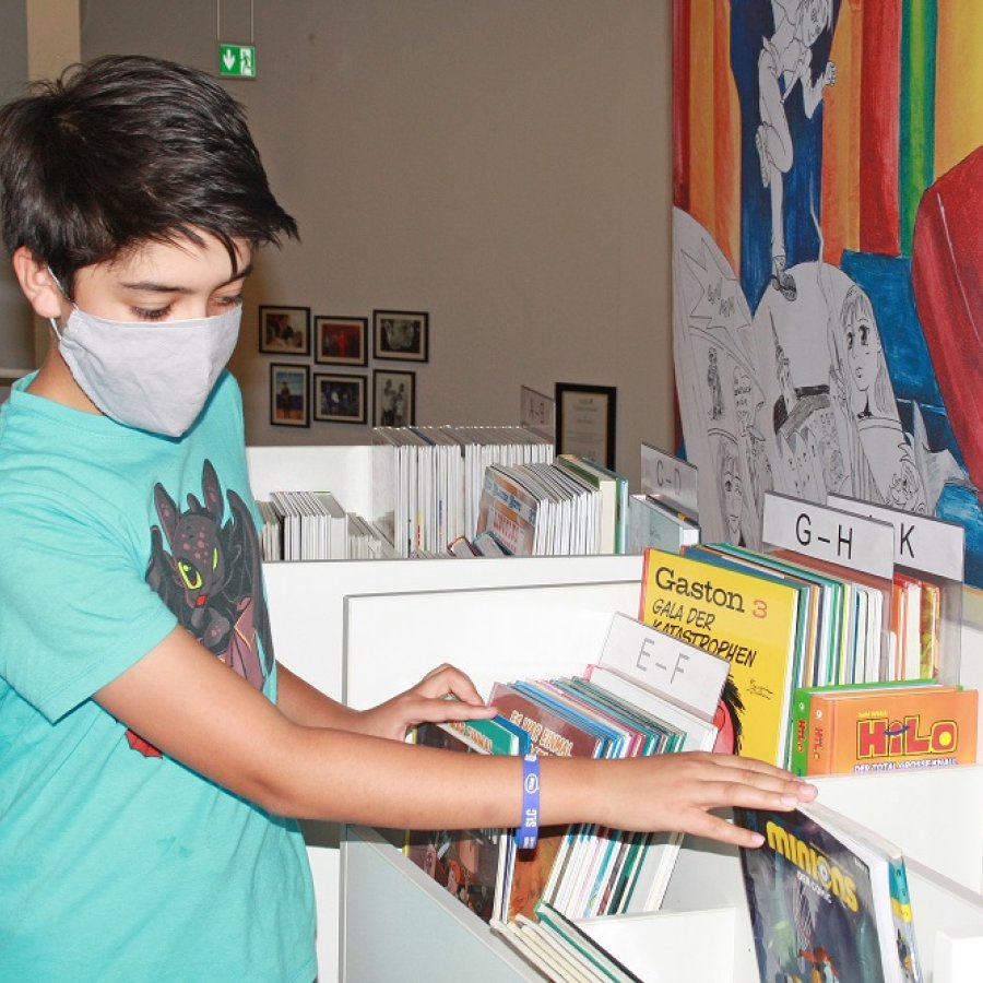 Junge mit Mundschutz blättert durch Comics in einem Regal
