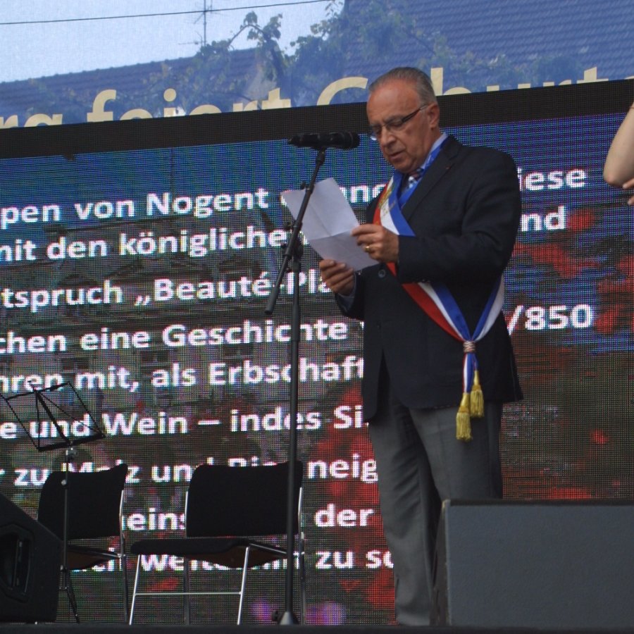 Mann mit Trikolore steht auf einer Bühne und liest eine Rede vom Zettel ab, die im Hintergrund auf digitaler Leinwand eingeblendet wird