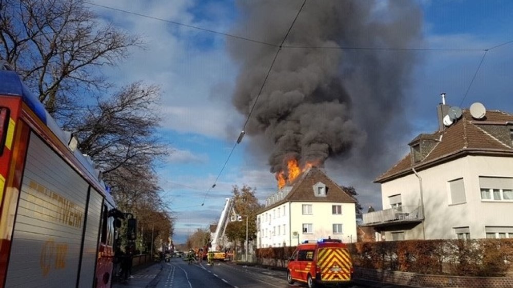 Feuerwehrautos vor einem haus mit brennendem Dachstuhl, Flammen und dunkler Rauch steigen auf