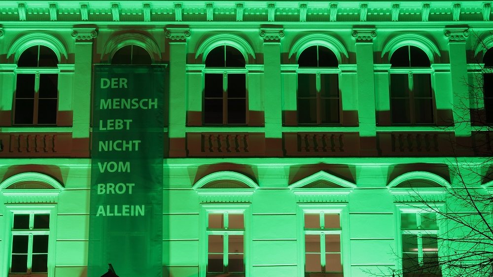 Banner mit der Aufschrift "Der Mensch lebt nicht vom Brot allein" und dem Hashtag "kultur" an der grün illuminierten Stadtmuseums-Fassade