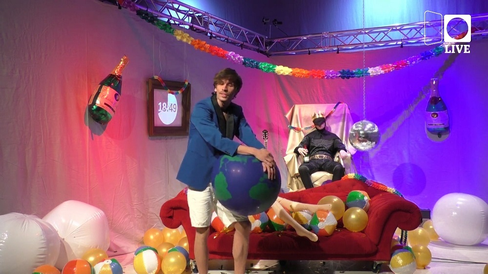Szene aus dem Theaterstück "Vorbei! Vorbei! - Die letzten 60 Minuten der Pandemie" - der Moderator mit einer aufblasbaren Erdkugel in der Hand steht vor einem roten Sofo mit Luftballons, im Hintergrund sitzt eine Fledermaus auf einem Thron.
