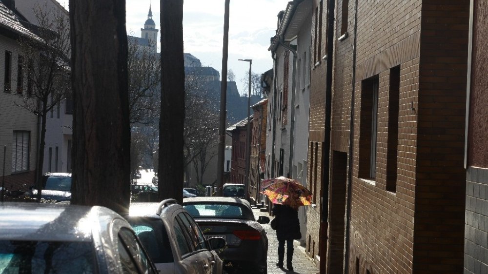 Person mit Regenschirm neben zwischen Häusern und Autos, in denen sich die Sonne spiegelt, mit Blick auf den Michaelsberg
