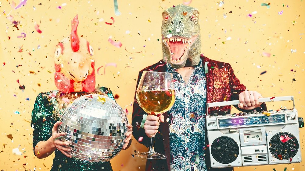 Schauspielr mit einer Hühner- und einer Dinosaurierkopfmaske, mit einer Partykugel, einem Sektglas und einer Stereoanlage in der Hand, hinter Konfettiregen