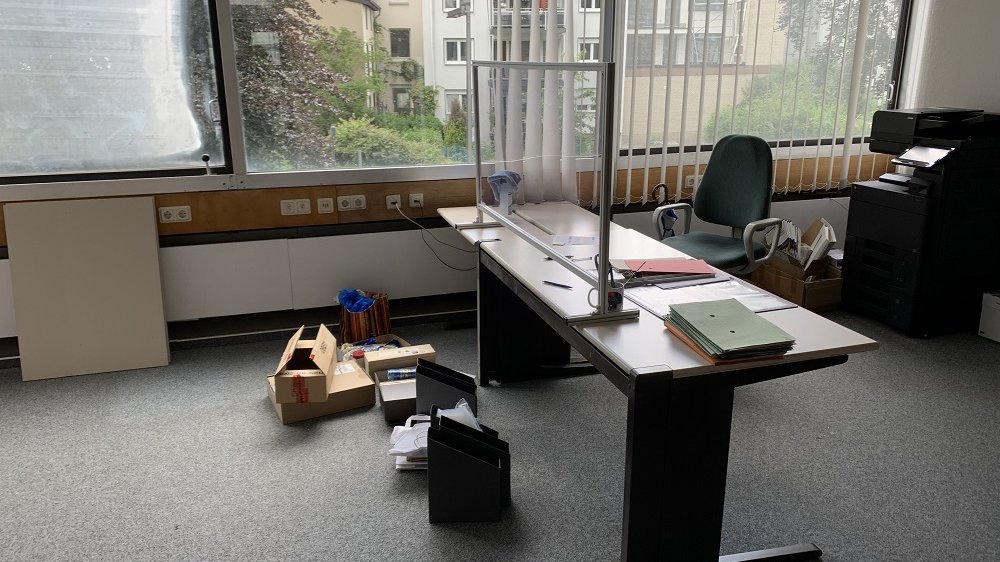 Tisch mit Spuckschutz, Kartons, ein Bürostuhl und ein Kopierer in einem ansonsten leeren Raum.