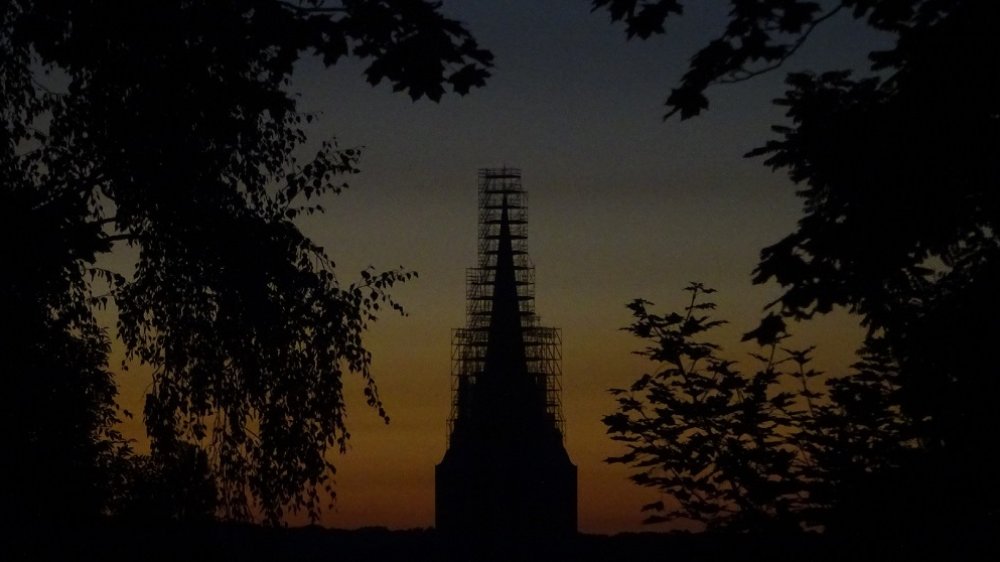 Silhouette des eingerüsteten Kirchturms von St. Anno vor dem Abendhimmel, eingerahmt von Baumsilhouetten.
