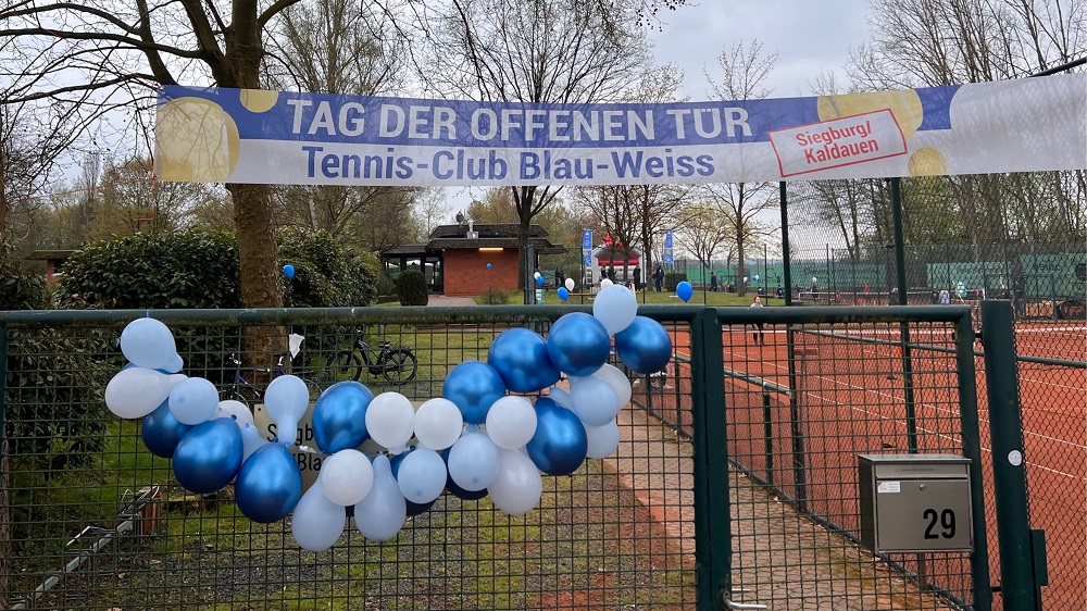 Banner "Tag der offenen Tür" und blau-weiße Luftballons an einem Zaun vor einem Tennisplatz.