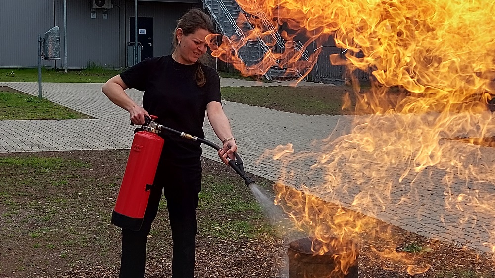 Frau mit Feuerlöscher an einem brennenden Fass.